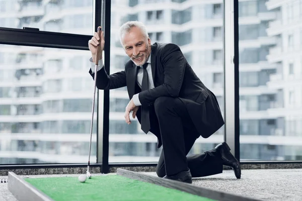 Уважаемый пожилой человек, играющий в мини-гольф в офисе. Он улыбается и смотрит на поле для мини-гольфа — стоковое фото