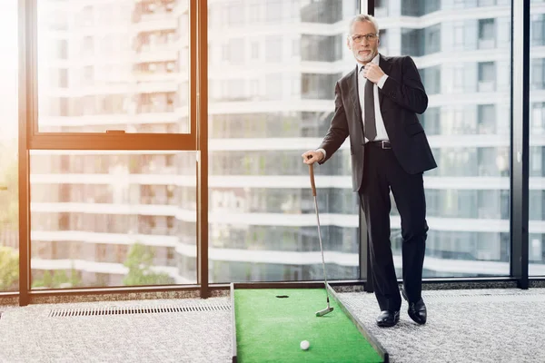 Un respetable anciano jugando un mini golf en la oficina. Se está preparando para atacar. — Foto de Stock