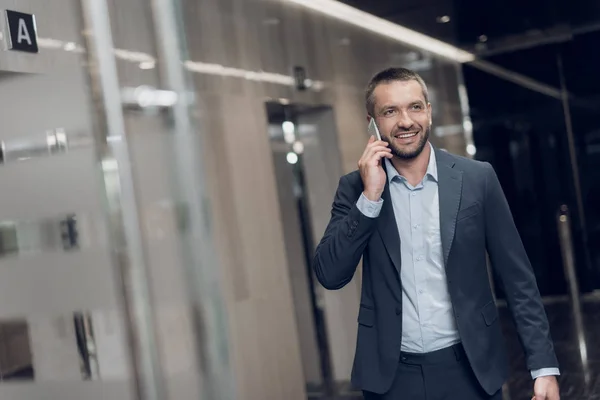 Уважаемый человек в строгом костюме ходит по офисному коридору с телефоном в руках, улыбается и разговаривает — стоковое фото