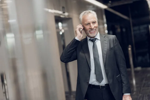 Un respetable anciano con un traje estricto camina por el pasillo de la oficina con un teléfono en las manos, sonríe y habla — Foto de Stock