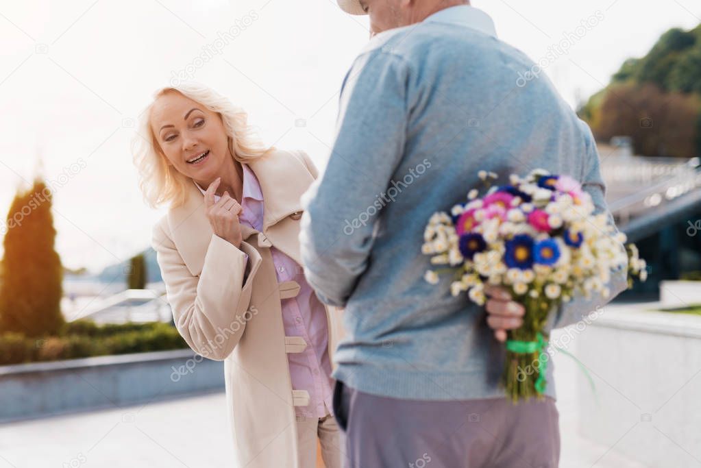 An elderly man is holding a bouquet of flowers behind back. A woman came on a date and tries to spy on what hides a man