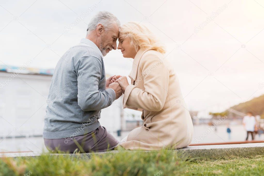 An elderly couple is sitting on the edge of a flower bed. They hold hands.