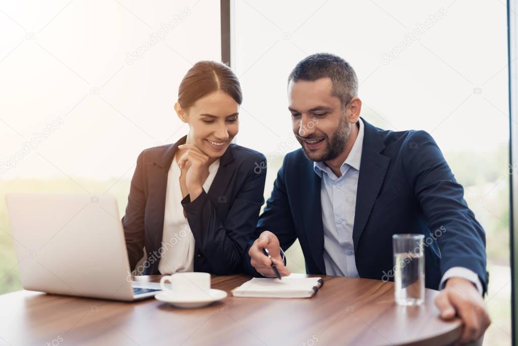 A man and a woman in strict business suits are discussing something while sitting at a table with a cup of coffee