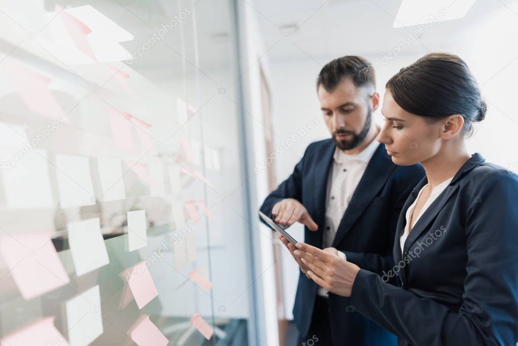 A man and a woman in strict business suits stand at a glass wall with paper stickers and look closely at them