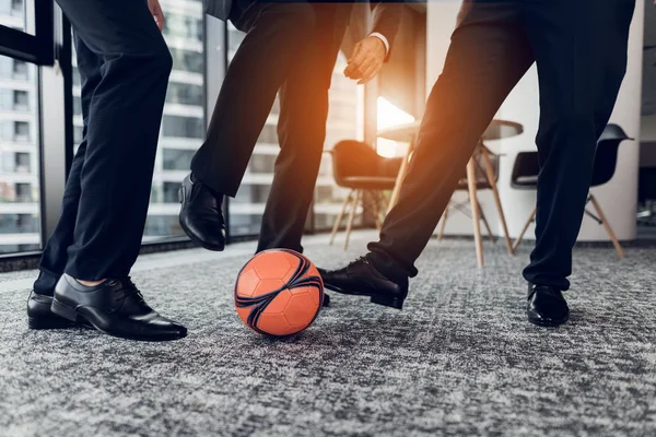 Закрывай. Трое мужчин в строгих брюках и черных туфлях играют в оранжевый мяч в футболе в офисе — стоковое фото