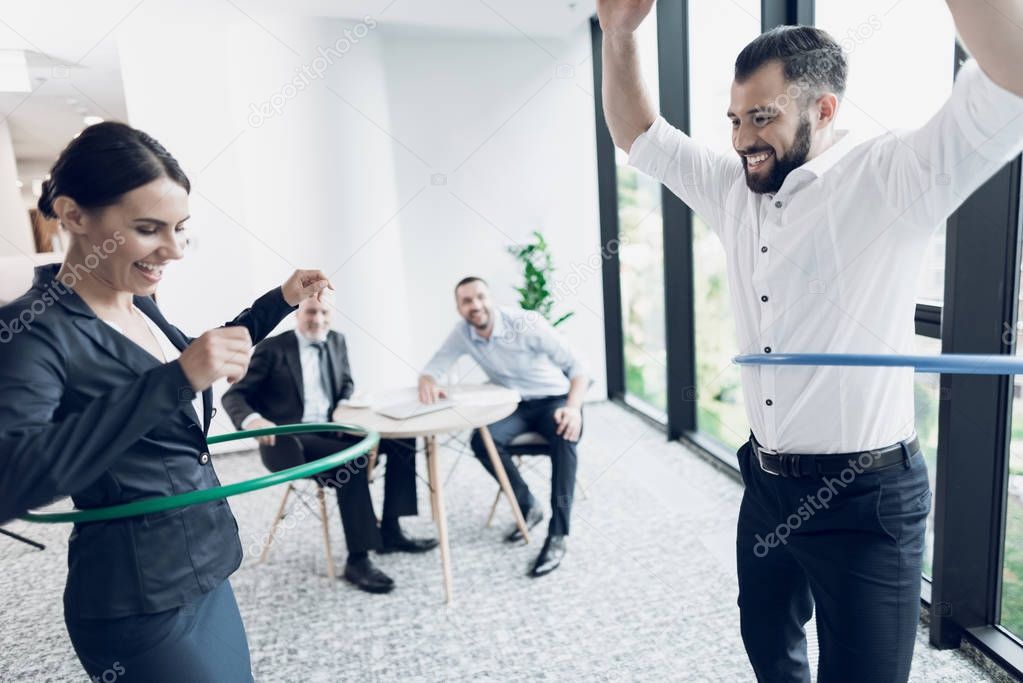 Fun in the office. A man and a woman twist hula hoops. Two of their colleagues sit at the table and look at them.