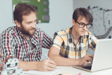 Bir adam ve bir genç bir dizüstü bilgisayar önünde bir masada oturuyor. Yakınlarda bir robottur. Onlar programı