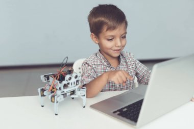 Yakın çekim. Küçük bir çocuk bir masada oturan ve gri bir dizüstü bilgisayar çalışma. Yakınlarda küçük bir robot olduğunu