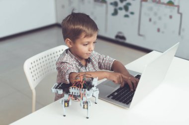 Yakın çekim. Küçük bir çocuk bir masada oturan ve gri bir dizüstü bilgisayar çalışma. Yakınlarda küçük bir robot olduğunu