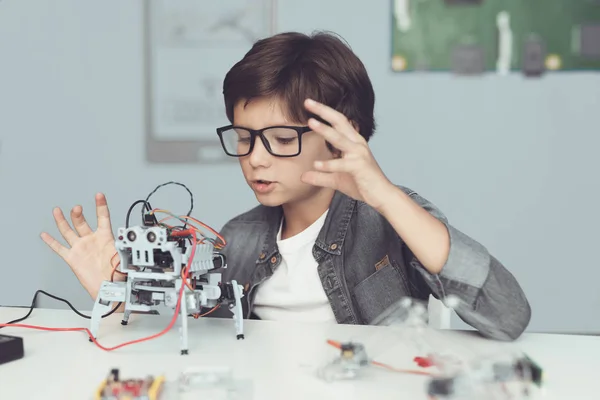 Ein kleiner Nerd mit Brille hält einen Roboter in der Hand. er schaut begeistert auf seine Schöpfung — Stockfoto