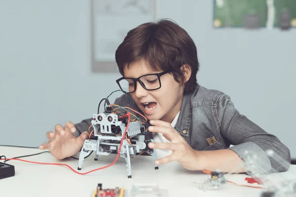 Een kleine nerd in glazen is het bezit van een robot. Hij kijkt enthousiast naar zijn schepping — Stockfoto