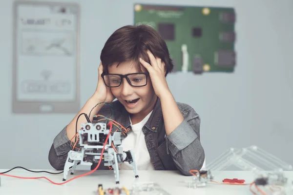 Ein kleiner Nerd mit Brille hält einen Roboter in der Hand. er schaut begeistert auf seine Schöpfung — Stockfoto