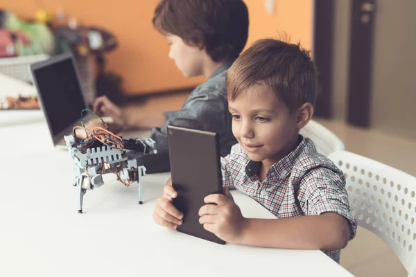 Dos chicos están sentados en el taller a la mesa. Uno de ellos se sienta detrás de un portátil, el segundo con una tableta . — Foto de Stock