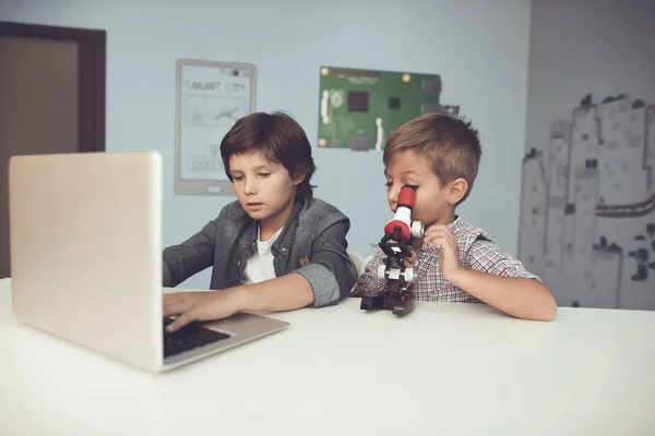 Two boys sitting at the table. One of them is sitting in front of a gray laptop. The second looks through a microscope — Stock Photo, Image