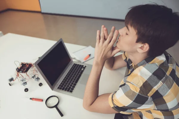 Un adolescente está sentado en una mesa frente a un portátil. Cerca hay un robot que él ensambló. — Foto de Stock