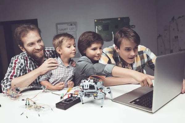 Two boys a teenager and an adult male are sitting in the workshop and constructing a robot — Stock Photo, Image