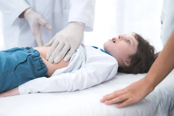 O médico de luvas brancas massageia a barriga de um menino doente . — Fotografia de Stock