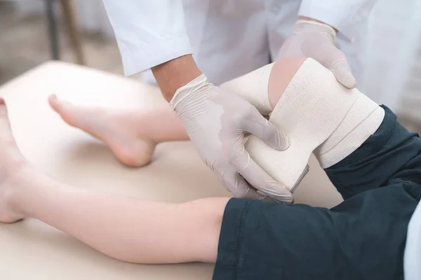 Dokter dalam sarung tangan putih memutar kaki anak laki-laki dengan perban . — Stok Foto