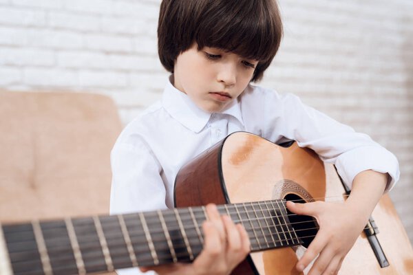 A little dark-haired boy in a white shirt is playing the guitar. The boy learns playing the guitar himself. He sits on the couch in a bright room.x