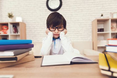 Oğlan ödevini oturuyor. Gözlüklü çocuk dalgın bir görünüme sahiptir ve masada oturuyor. Masada ders kitapları ve egzersiz kitaplar vardır.