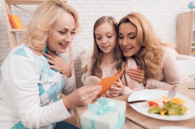 İki yetişkin kadın ve bir kız bir tebrik kartı arıyoruz. Bu annem ve büyükannem kızın. Evde yemek var. Onların masaya sebze ve tatlılar ellerinde.