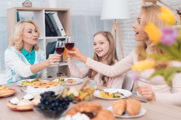 Mom and grandmother are drinking wine, a little girl is drinking juice. They have dinner at home. They have vegetables and sweets on their table.