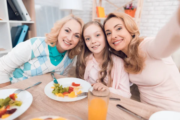 Mom, grandmother and granddaughter do selfie at the dinner table. They have dinner at home. They have vegetables and sweets on their table.