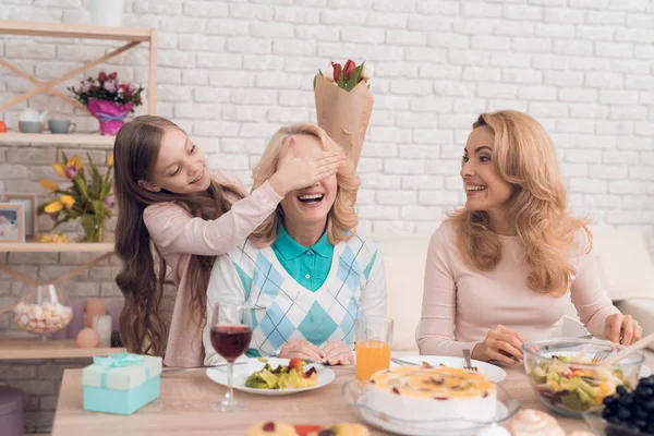 The girl closes eyes to her grandmother. She wants to make a surprise. They have dinner at home. They have vegetables and sweets on their table.