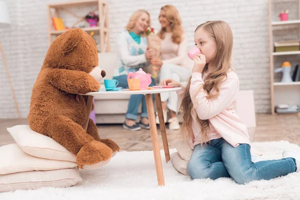 小女孩在玩一只玩具熊 玩具桌上有玩具杯 坐在她的母亲和祖母的身后 — 图库照片