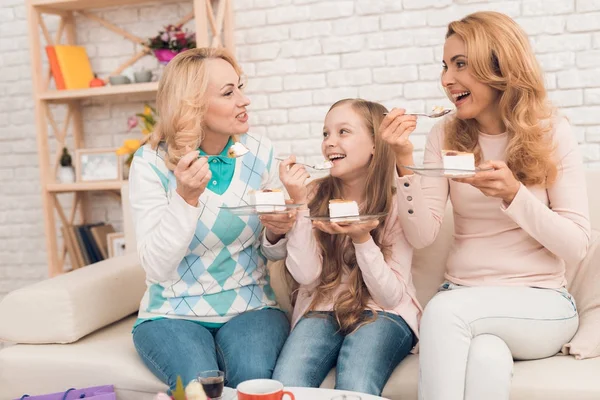 Mom, grandmother and little girl eat cake on the couch. They have a good mood, they smile at each other.