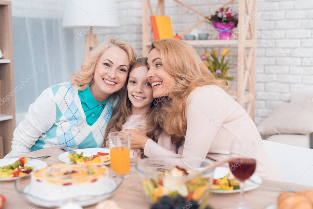 Two adult women and a girl are hugging, sitting at a table. This is the mother and grandmother of the girl. They have dinner at home.