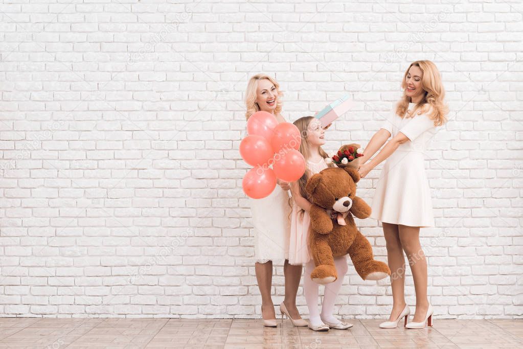 Mom, grandmother and girl are posing together near a light wall. They keep flowers, balloons and a toy bear in their hands.