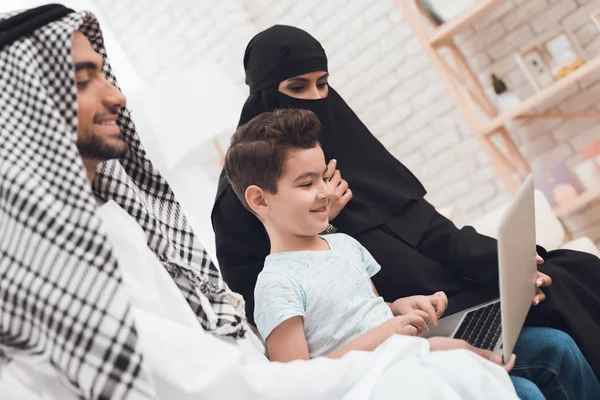 A traditional muslim family at home looks at the laptop screen. On the couch sat mother, father and little boy. The boys mother wears a parandja.