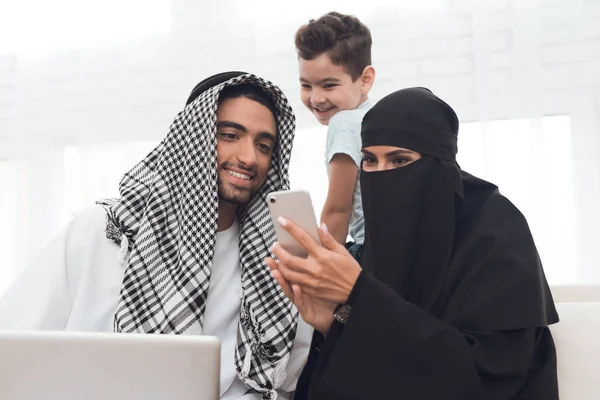 一名身穿布卡的阿拉伯妇女正在看手机 一个男孩和一个成年男子坐在一起 这是一个传统的穆斯林家庭 — 图库照片