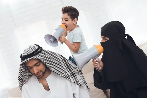 A woman in a burqa and a boy scream at a man in traditional muslim clothes. In their hands they hold speakerphones. A man has a laptop in his hands.