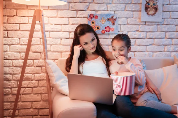 母亲和小女儿看电影在笔记本电脑和吃爆米花在晚上在家 — 图库照片