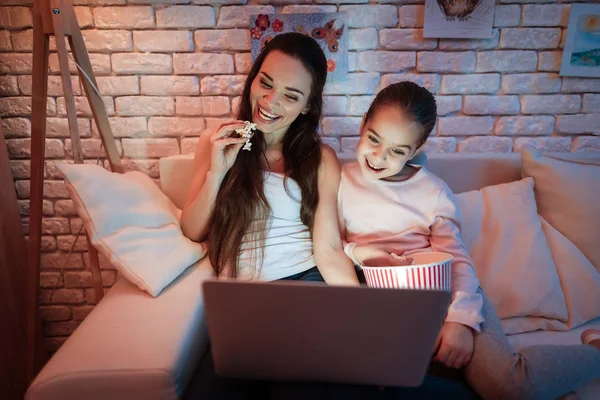 母亲和小女儿看电影在笔记本电脑和吃爆米花下灯罩在晚上在家 — 图库照片