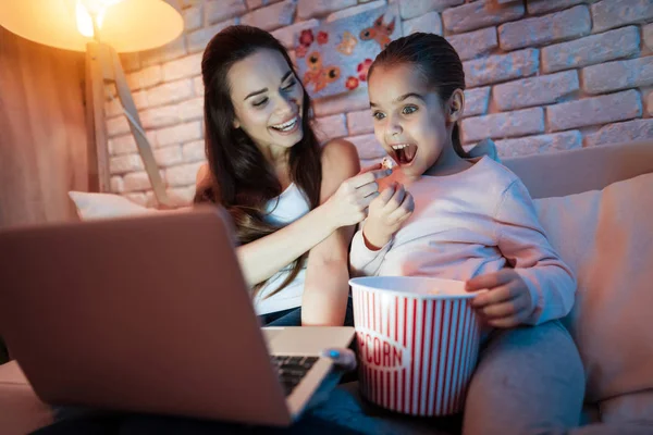 母亲和小女儿看电影在笔记本电脑和吃爆米花下灯罩在晚上在家 — 图库照片