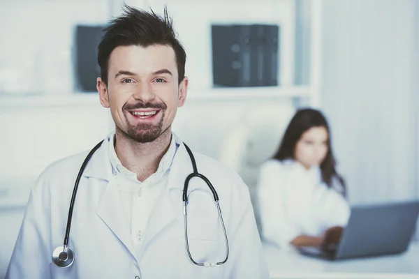 Χαμογελαστά αρσενικό ο γιατρός βλέπει η κάμερα. Βοηθός κάθεται στο τραπέζι με φορητό υπολογιστή — Φωτογραφία Αρχείου