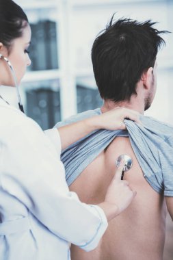 Genç doktor steteskopla hastanın akciğerlerini inceliyor.
