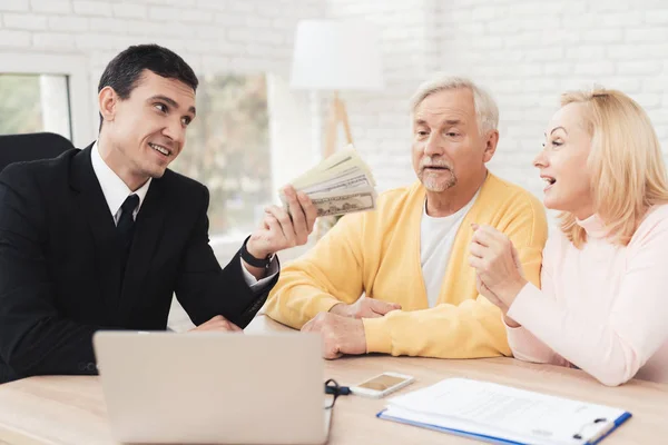 Mature couple at a reception with a lawyer. The lawyer holds a bundle of money and smiles. Old people sit next to him and look at him. They are sitting at the desk in the lawyer's office.