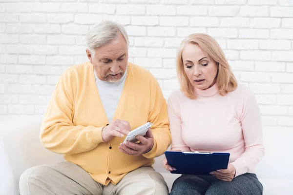 A couple of old people are sitting on the couch and are concentrating something. A man has a yellow cardigan, a pink sweater on a woman. They have documents and a calculator in their hands.
