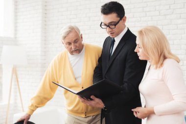 Bir emlakçı ile bir resepsiyonda eski insanların bir çift. Bir emlakçı yanındaki stand ve şok edici emlakçının klasöründeki formunu okuyun. Emlakçı siyah bir takım elbise içinde yan yana duruyor.