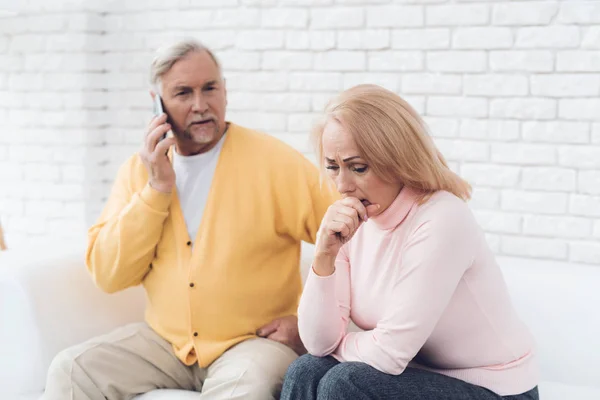 A man in a yellow cardigan is talking on his smartphone. next to his wife sits and worries. She is very worried. They are sitting on a white sofa in their living room.