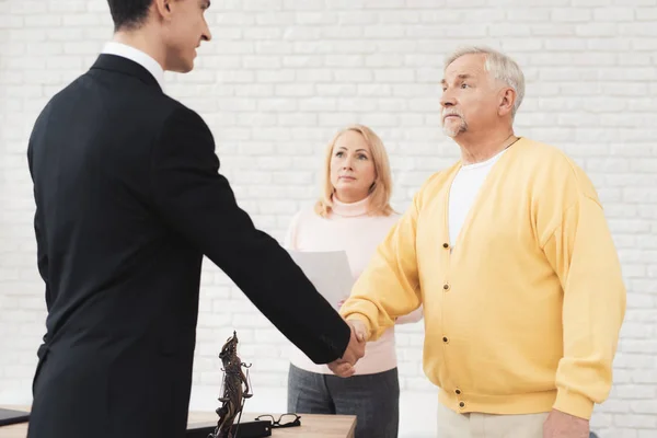 A couple of old people came to see a realtor. An old man in a yellow cardigan greets a realtor in a black suit. Behind him stands a woman in a pink sweater.