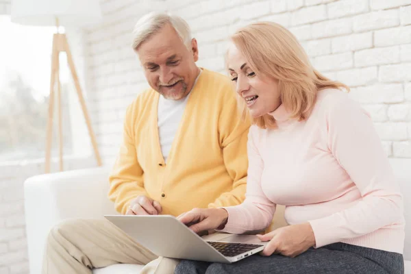 A pair of old people are sitting in front of a laptop. They found the home of their dreams and are happy about it. They are sitting on a large white sofa in their apartment.