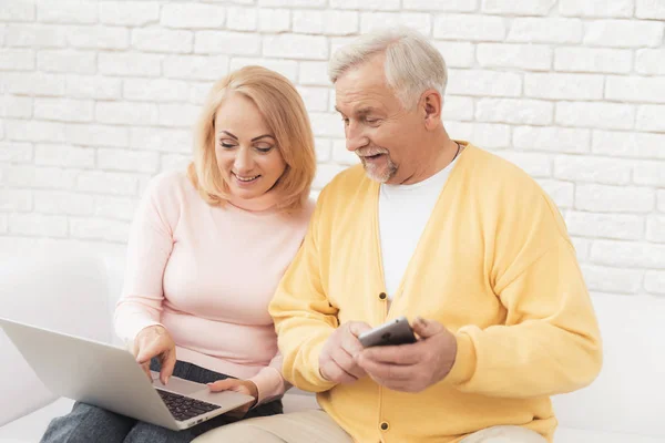 一个年长的男人和女人坐在一个白色的大沙发上 看着笔记本电脑屏幕上的东西 他手里的那个人有一把智能手机 他们坐在一间宽敞明亮的起居室里 — 图库照片