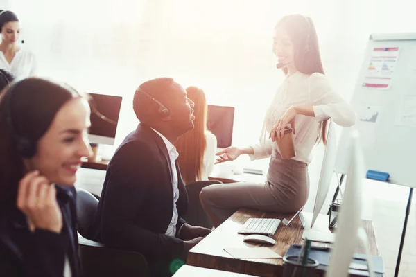 Ein chinesisches Mädchen sitzt auf dem Tisch im Callcenter und spricht mit einem Schwarzen. — Stockfoto