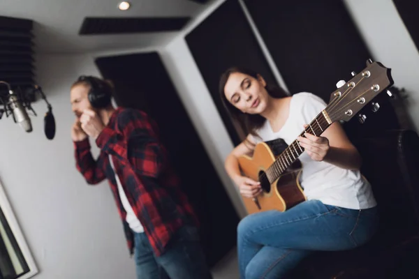 A man and a woman sing a song at a recording studio. A man sings, and a woman plays the guitar.
