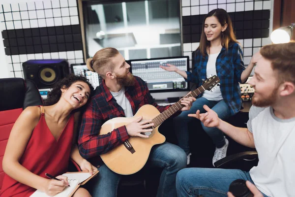 Two women and two men sing a song in a guitar in a modern recording studio. They sit behind the remote control of sound and sing a song. They are having fun.